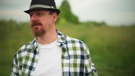 close-up of an artist wearing a hat and checkered shirt, standing still in a lush grassland. the artist, holding a palette, gazes thoughtfully at the surrounding nature