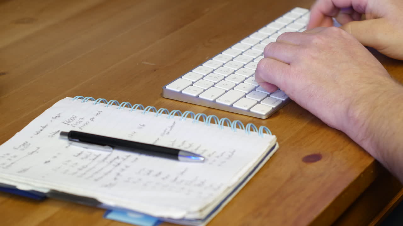 Man working on computer typing on a keyboard on a desk and taking notes on a notepad with a pen