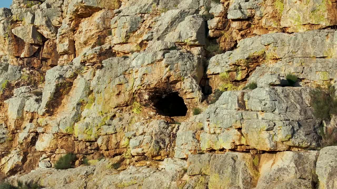 70mm reverse drone flight from inside a cave on an impressive rocky hill. As the camera zooms out, vivid colors and plants appear, revealing the scale of the formation under a blue sky