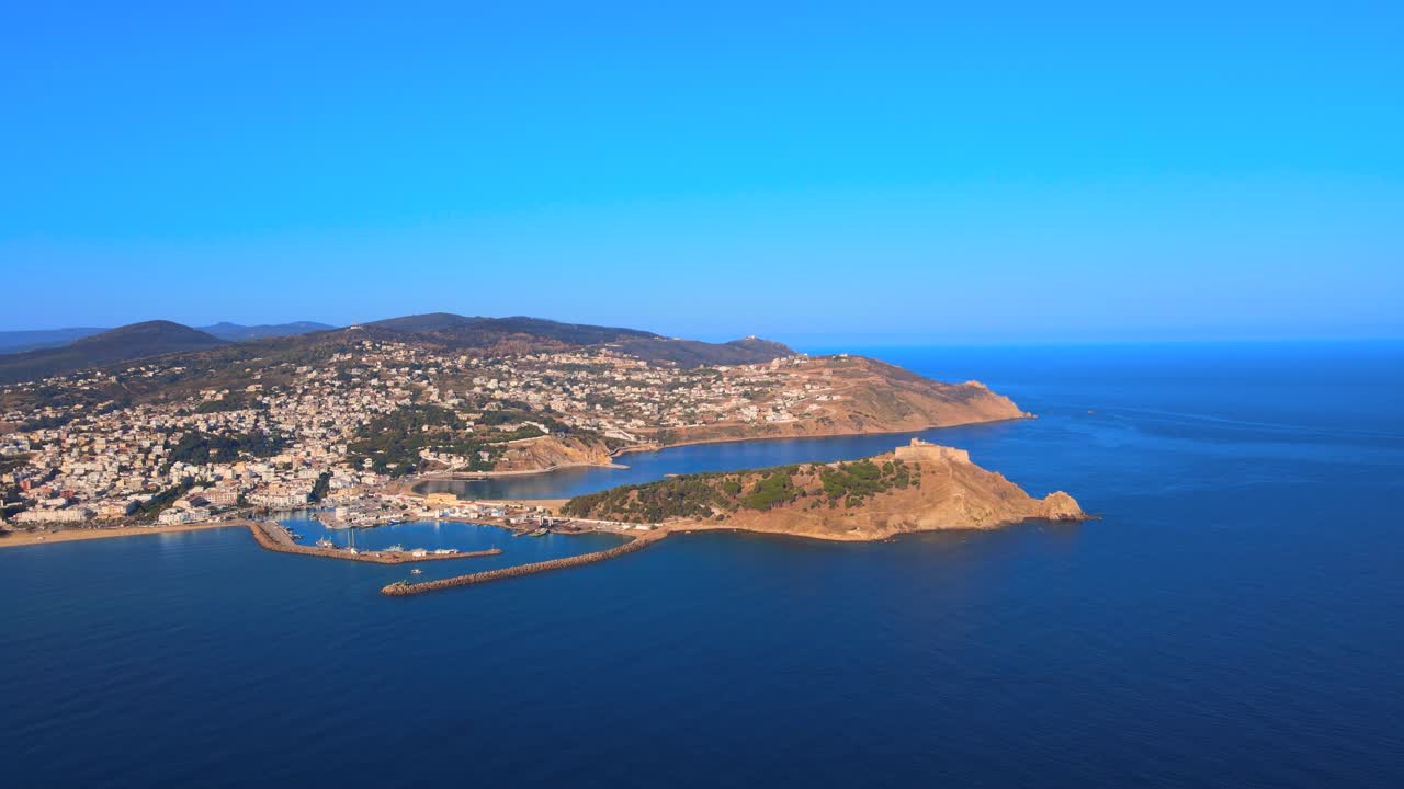 An aerial view of water and land on the coast in North Africa. The ocean is full of boats and an island can be seen in the distance. A beautiful city overlooks the water and mountains.