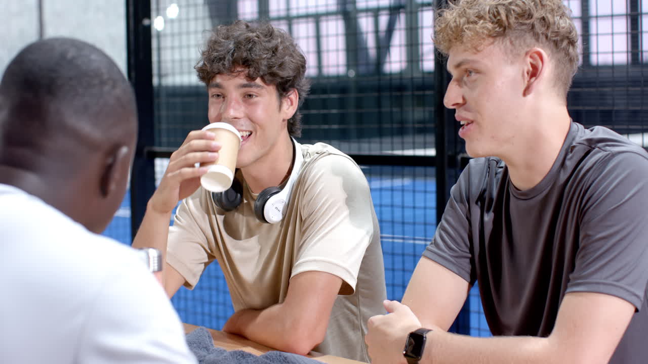 Young diverse men enjoying conversation and coffee after playing padel tennis on indoor court