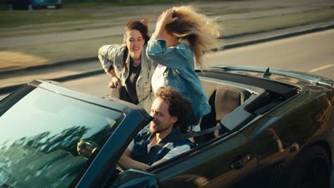 a happy trio: a guy and two girls are riding in a gray convertible. a blonde girl and a brunette girl have fun and wave their hands and look at the camera