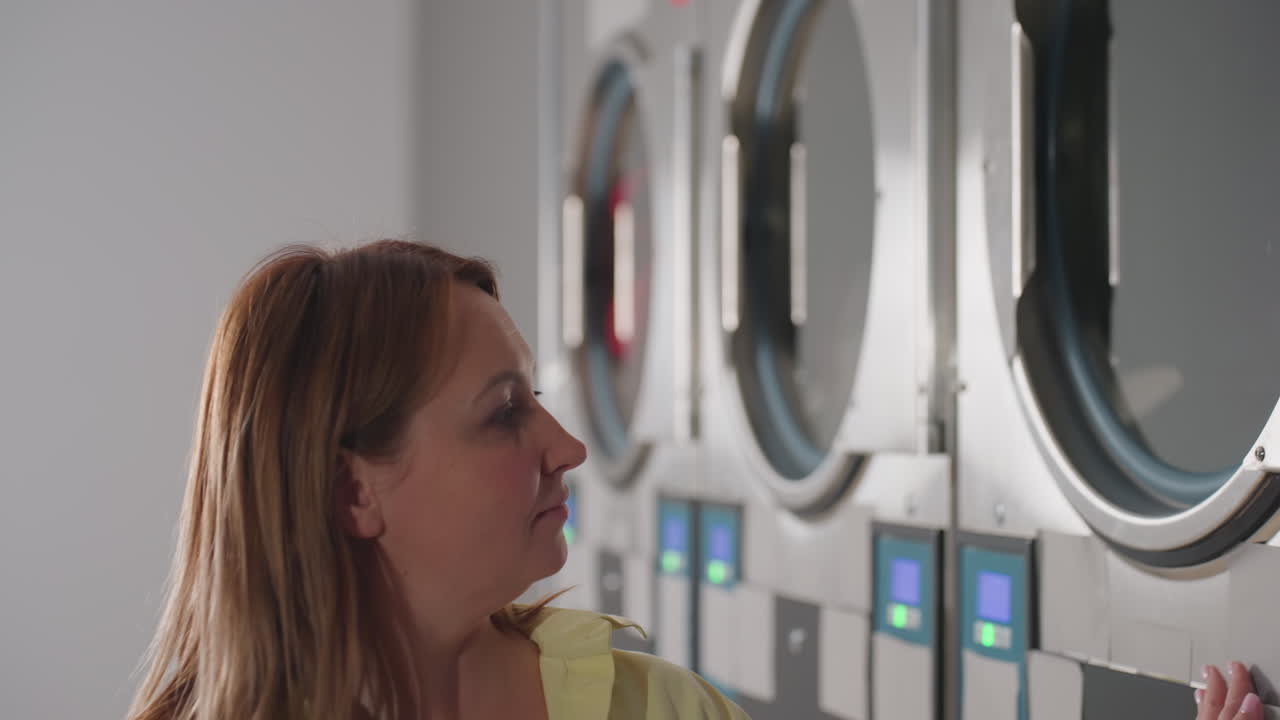 Zoom in on housewife glides hand across industrial washer while clothes tumble inside, digital controls glowing, stainless drums humming in modern laundromat, showcasing service, hygiene, reliability