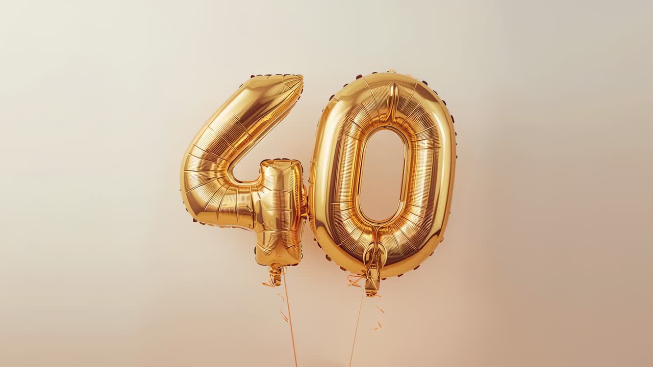 Swaying from gentle air current, metallic gold '4' and '0' balloons rotating in studio with ribbons