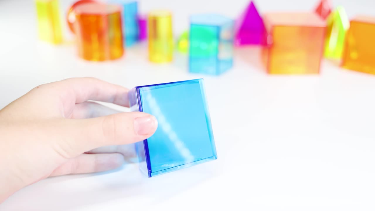 Hands manipulate colorful 3D geometric shapes on a white surface, highlighting educational exploration and tactile learning in bright lighting