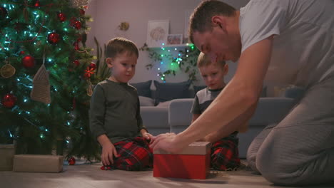 Vater-Mit-Zwei-Kindern-öffnet-Ein-Weihnachtsgeschenk-Unter-Dem-Baum-Und-Freut-Sich.-Ein-Geschenk-Für-Meinen-Vater.-Ein-Lichtstrahl-Des-Geschenks-Scheint-Auf-Das-Gesicht.-Freuen-Sie-Sich-Mit-Den-Kindern-über-Das-Neujahrsgeschenk.-Gemeinsam-Ein-Geschenk-öffnen.-Hochwertiges-4K-Filmmaterial