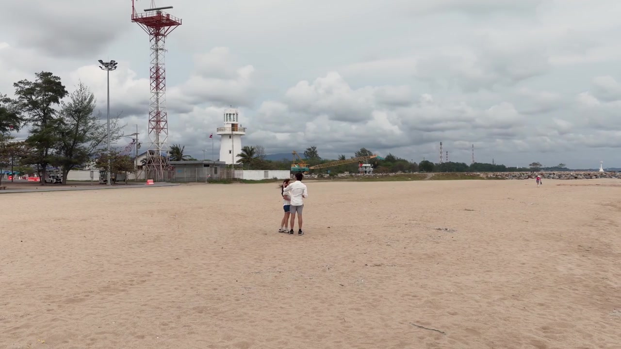 A câmera recua revelando um jovem casal em pé em uma extensa praia de areia na Tailândia