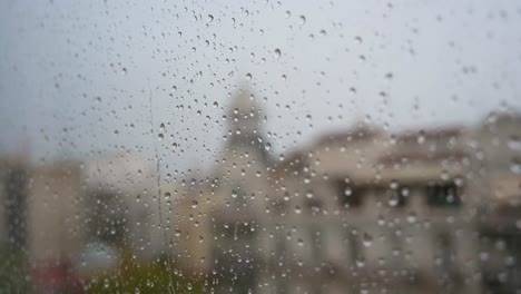 Una-Vista-Cercana-En-Cámara-Lenta-De-Fuertes-Gotas-De-Lluvia-Vistas-A-Través-De-Una-Ventana,-Con-Un-Paisaje-Urbano-Al-Fondo