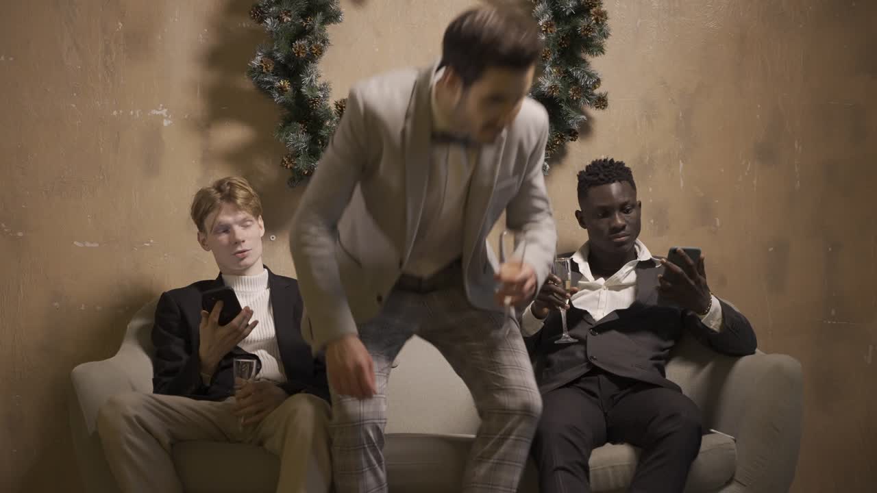 Two friends are sitting on the couch using mobile and drinking champagne at New Year's party, another friend sits and they start talking