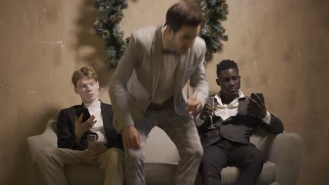 Two friends are sitting on the couch using mobile and drinking champagne at New Year&#039;s party, another friend sits and they start talking