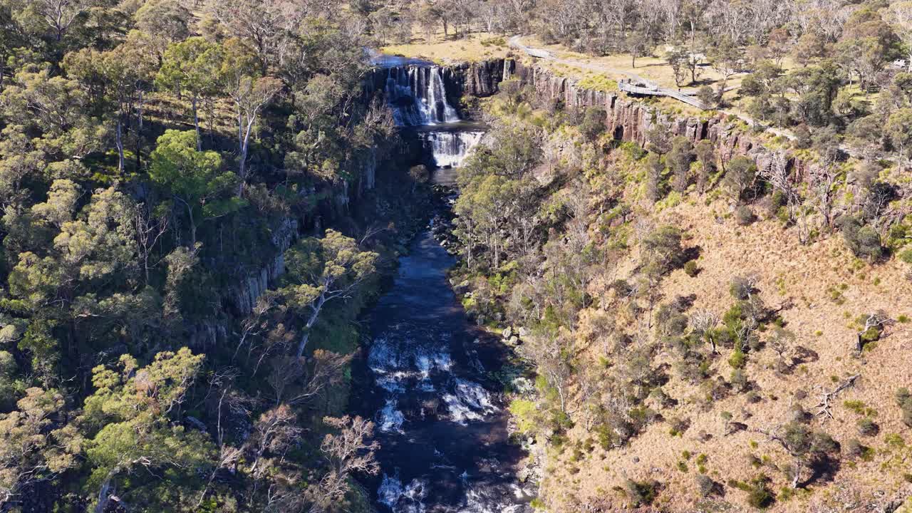 Drone camera smoothly descends above Ebor Waterfall, revealing cascading water, rugged cliffs, and dense forest in bright natural daylight