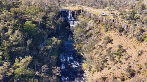drone camera smoothly descends above ebor waterfall, revealing cascading water, rugged cliffs, and dense forest in bright natural daylight
