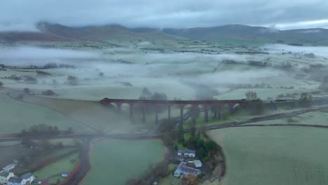 Derelict-stone-viaduct-bridge-slow-orbit-showing-mist-and-fog-with-distant-low-mountains-at-dawn-in-winter