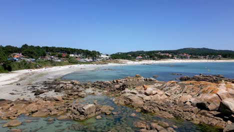 Aerial-Footage-of-Silent-Beach-Close-to-Forest-and-Houses-over-Rocky-Sunny-Shore