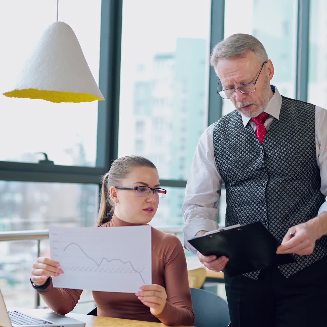 Elderly businessman and a secretary in the office. Attractive company worker sitting at the desk and talking to her boss about some graphics.