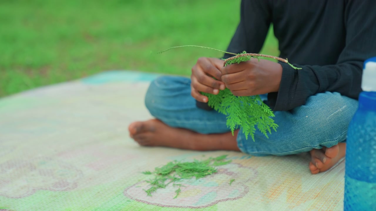 Wide shot child sitting on mat in grass holding branch, hands picking small leaves with steady focus, casual barefoot posture, relaxed outdoor mood, little forager practicing craft and learning
