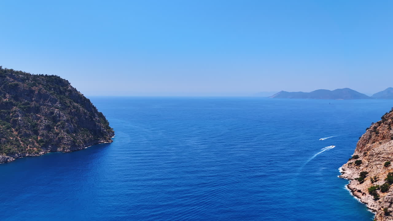 Flying above the spectacular blue of the Mediterranean Sea. Cliff rocks stick out of water. Some boats move by the waterscape. Turkey