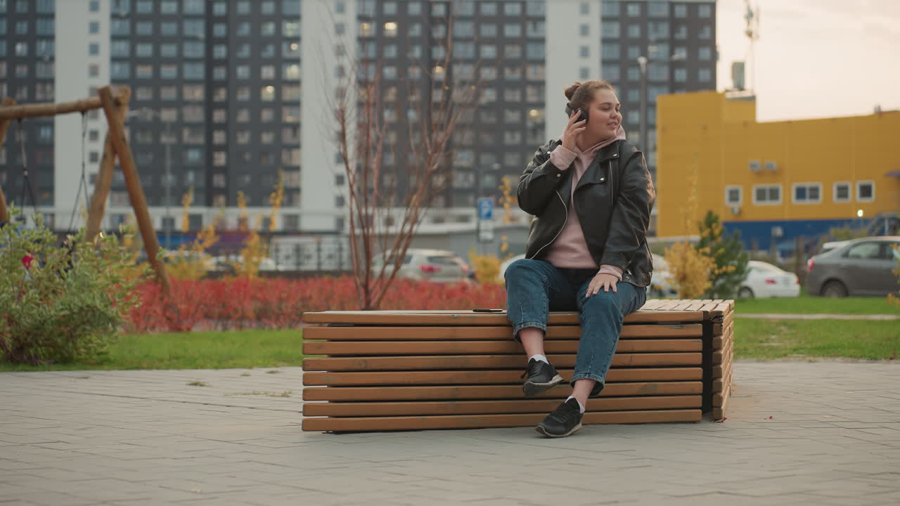 Young woman seated on wooden bench outdoors wearing pink hoodie listens to music with headphones while looking around surrounded by colorful plants parked cars and tall office buildings