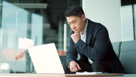 portrait thoughtful asian businessman working on a laptop computer at a modern office desk. confident focused pensive man in formal suit indoors. thinking of inspiration solving a problem.