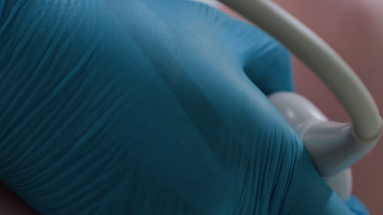 Close up of female lab technician wearing blue gloves gently examining patient throat area using ultrasound transducer during diagnostic procedure with careful precision in clinical environment