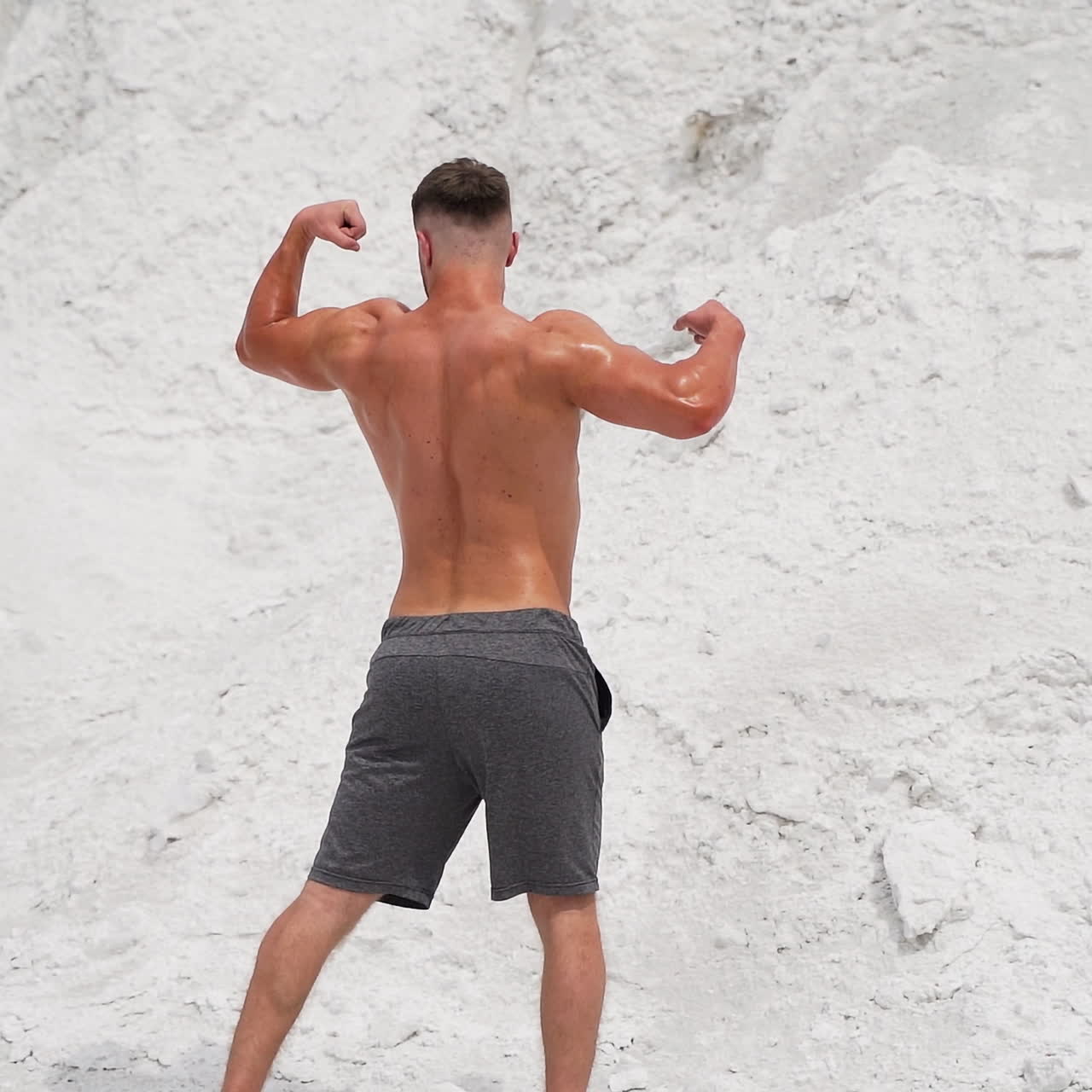 Backside view of a bodybuilder outdoors. Strong athlete without shirt shows his muscular body on white mountain background. Slow motion.