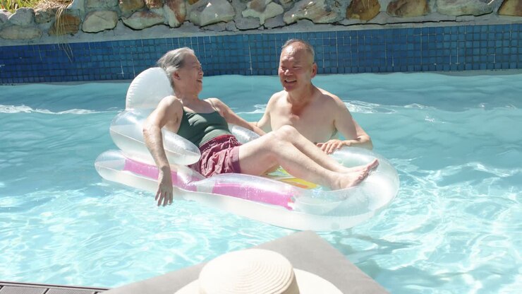 Diverse senior couple talking after woman shifting legs, man steadying float to help at pool