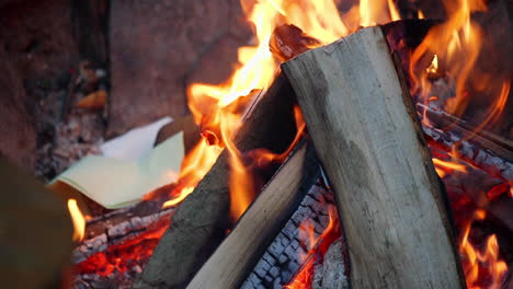 static close up fhd shot of papers and logs burning in an outdoor fire at a campsite with hot charred coals below and flames all around