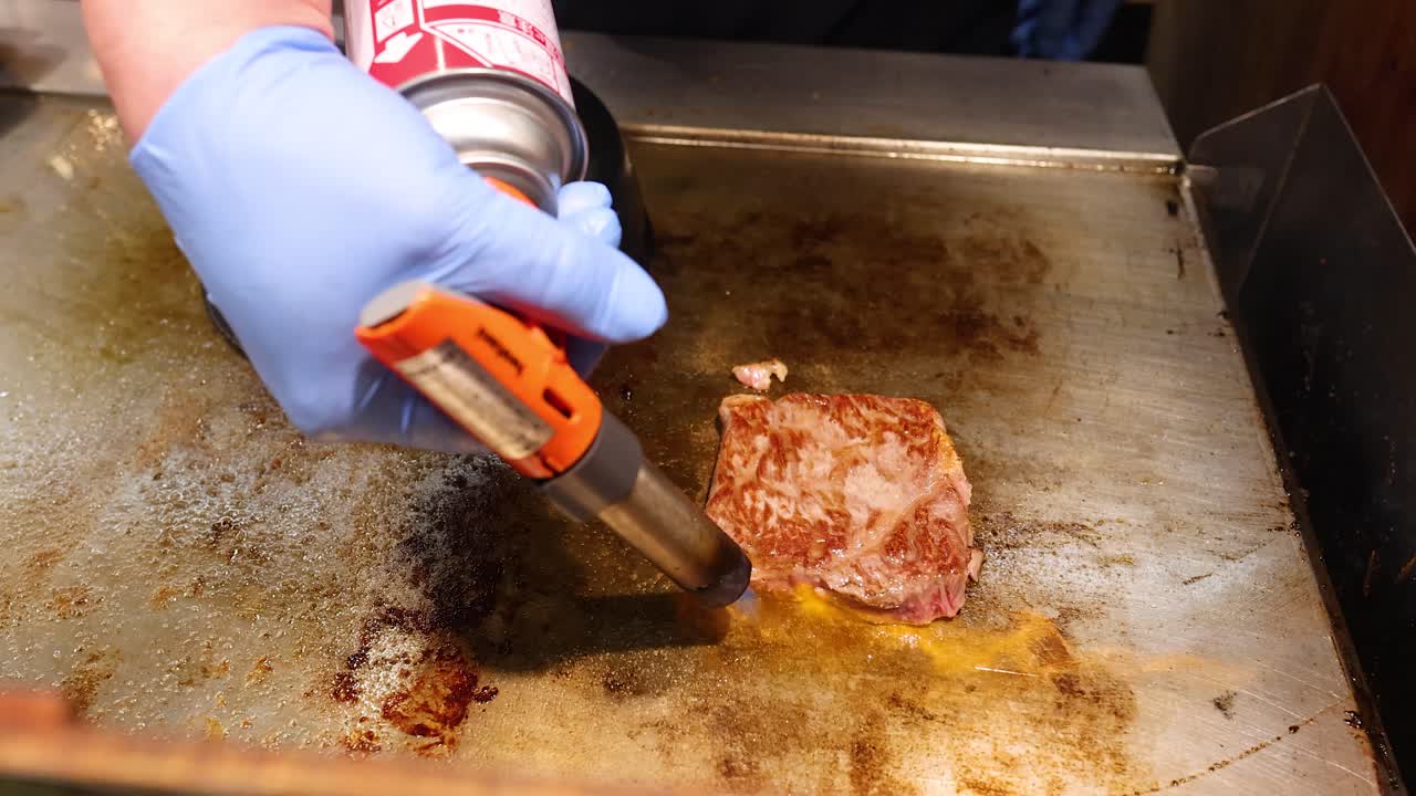 A chef uses a blowtorch to sear a wagyu beef steak on a flat grill