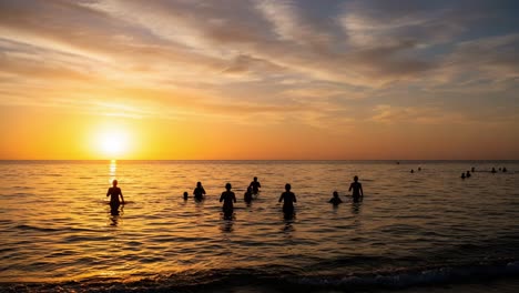 a tranquil sunset scene capturing silhouettes of people wading in the calm water, with vibrant hues of orange and gold filling the sky as day transitions to night