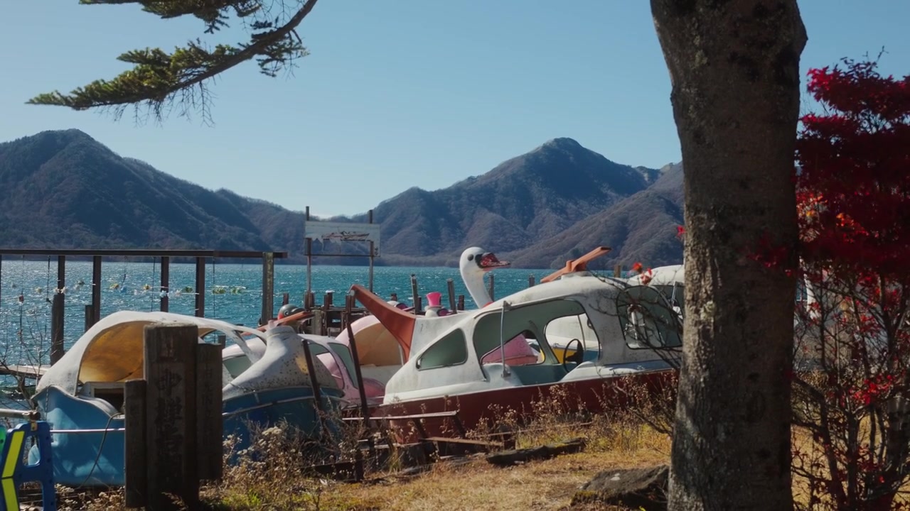 柔らかな日差しの中、湖畔にカラフルなボートが停泊し、背景には山々があり、木々に囲まれている。