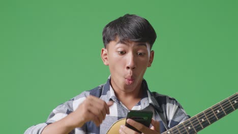 close up of asian teenager with guitar using smartphone and saying wow while standing on green screen background