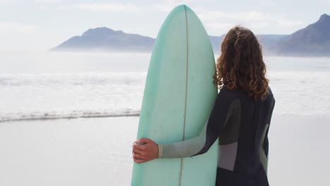 rear view of mixed race woman standing on beach holding surfboard looking out to sea