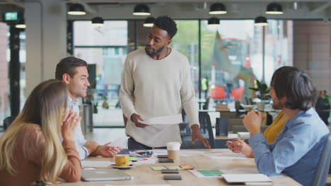 Businessman-Giving-Presentation-To-Colleagues-Sitting-Around-Table-In-Modern-Open-Plan-Office