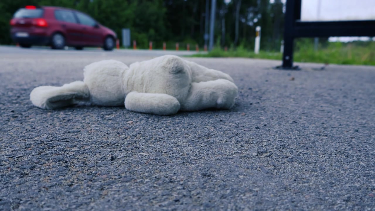 Coniglio di peluche solitario e abbandonato sdraiato sul ciglio della strada asfaltata mentre le auto passano sullo sfondo sfocato, a simboleggiare l'infanzia perduta, la solitudine, il pericolo e i ricordi dimenticati