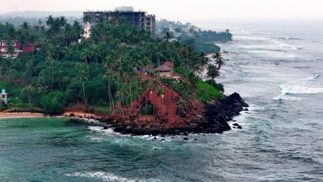 sri lanka's hazy coast visitors scale a red-earth headland amidst green palms dark rocks, overlooking a choppy grey-blue sea elevated wide shot reveals