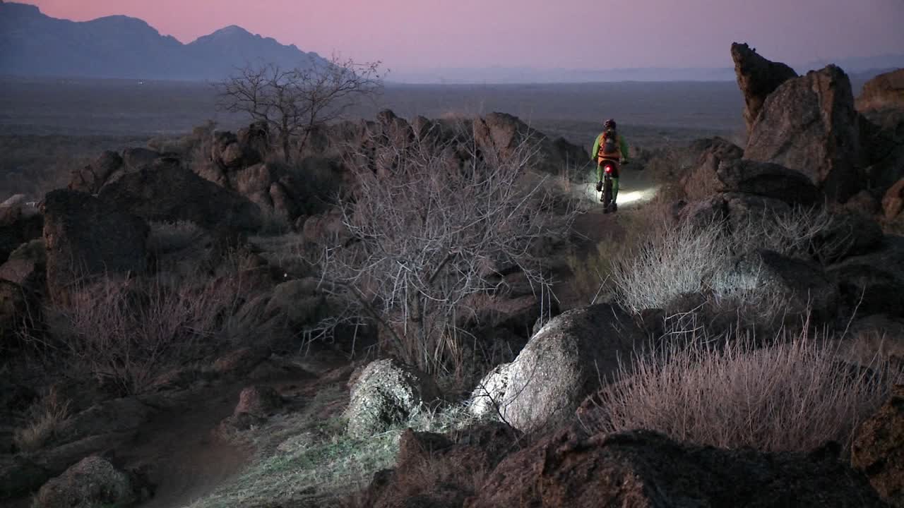 자전거 타는 사람은 헤드 라이트와 함께 황혼 근처의 사막 지역에서 거친 길을 여행합니다.