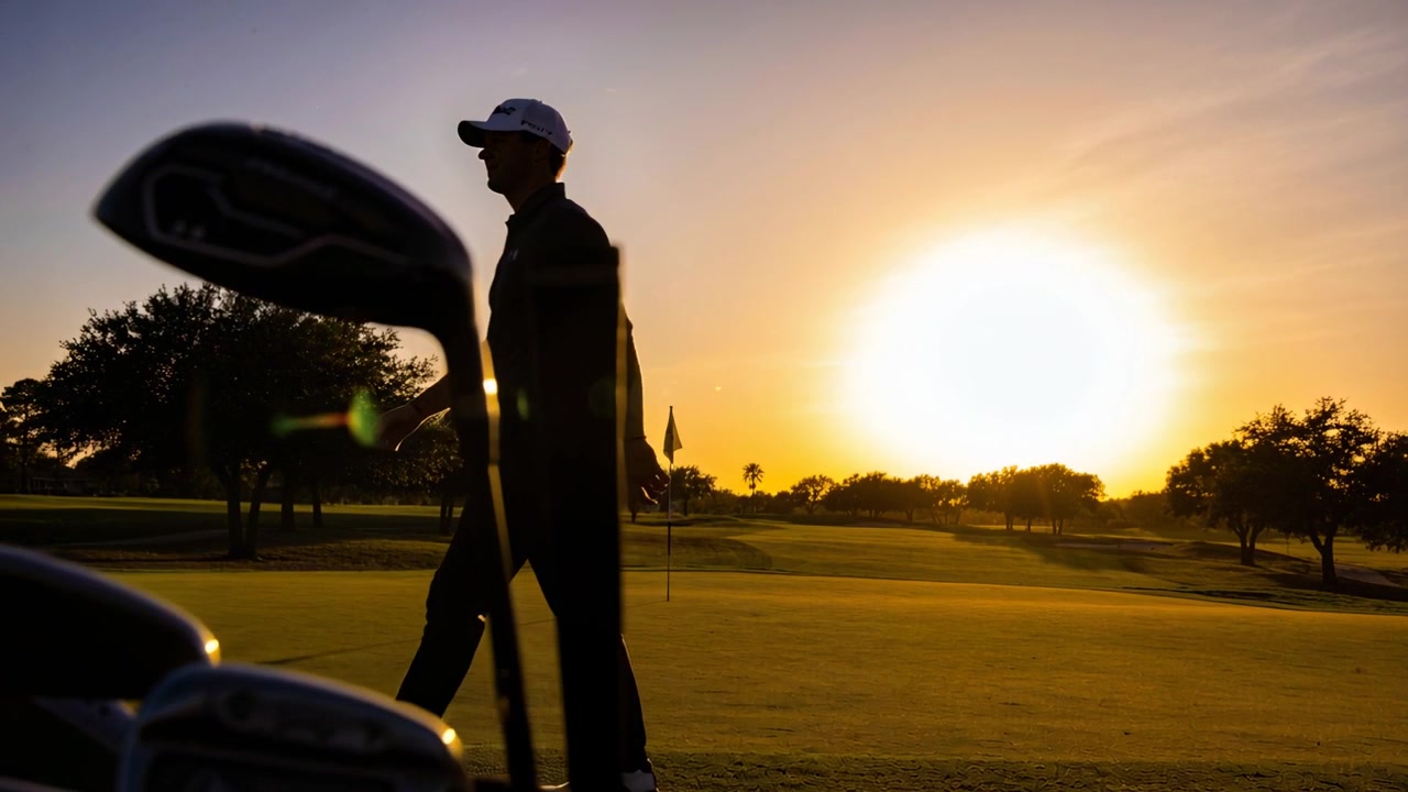 Mannelijke golfer met poloshirt en pet loopt over de green richting de vlag, langs een van achteren verlichte golftas