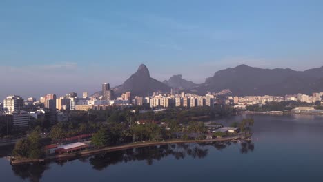 inclinación aérea hacia arriba que muestra la isla del lago de la ciudad y el club caiçaras en ipanema en primer plano, revelando leblon y la montaña de dos hermanos en el fondo al amanecer temprano en la mañana