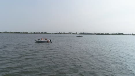 drone slomo of small boat on river with male friends swimming and hanging on the boat, with passing boat in background