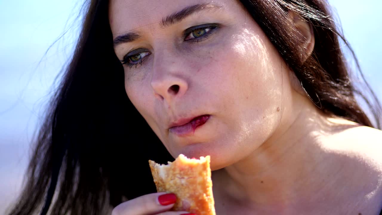 sea beach. portrait of beautiful woman, with long dark hair, with apetite eating cherry jam pie with tea. picnic outdoors, best breakfast