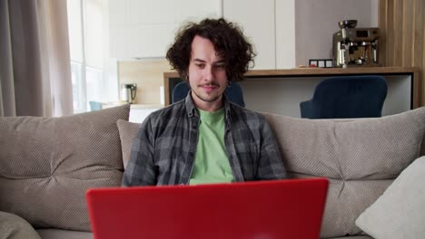 Focused-happy-brunette-guy-with-curly-hair-in-a-gray-checkered-shirt-sits-on-the-sofa-and-works-at-his-red-laptop-in-a-modern-studio-apartment