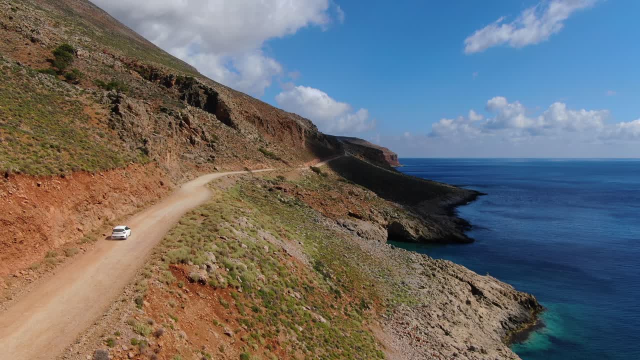 Crete Greece a white car drives to Balos Beach on a seaside cliff, Aerial follow shot with blurred plates pan-right