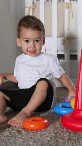 cute toddler boy playing with toy pyramid. domestic black cat lies on the floor near the kid. vertical video