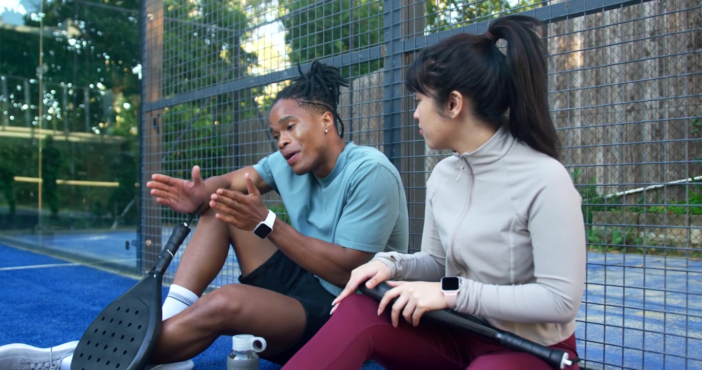 Diverse training partners sitting on blue court edge after play, discussing strategy with paddles