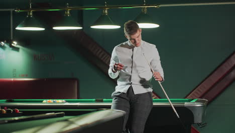 chap in white shirt and grey trousers sits on green pool table, holding cue stick and carefully applying red cover with a focused expression. another pool table is visible in the background