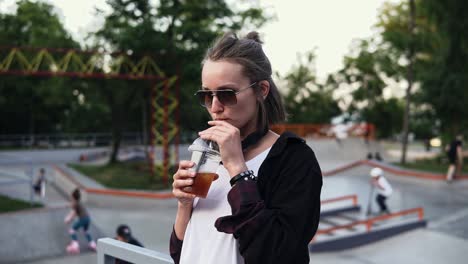 A-stylish-girl-in-sunglasses-and-headphones-enjoys-a-drink-in-a-transparent-glass,-drinks-through-a-straw.-A-woman-stands-in-the