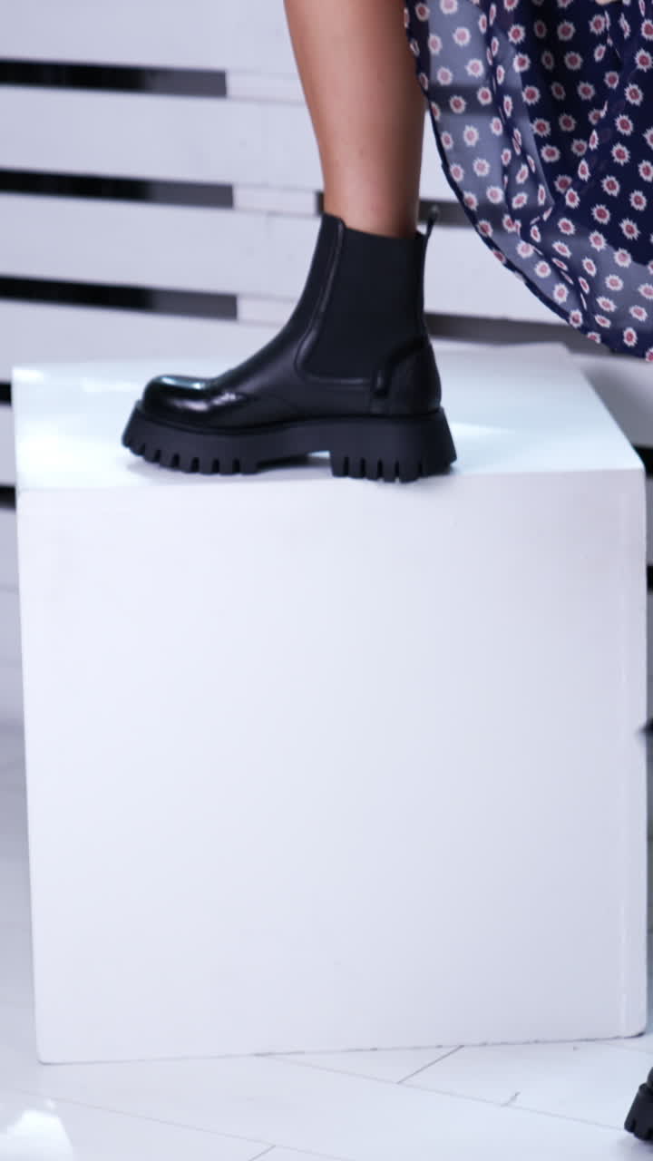 Female in short skirt and black leather boots with black bag. Lady puts her foot on the white cube to demonstrate her modern footwear. Vertical video