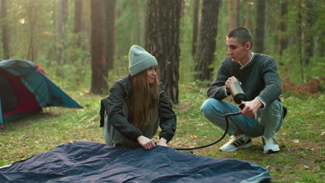 lady gazes flirtatiously at man pumping air into tent bag during outdoor moment in natural campsite, expressing lighthearted attraction with soft smile and warm body language
