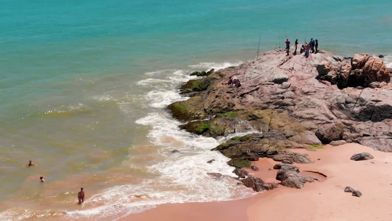 Static drone showing the shore of Itapuã while at the top of the rock some fishermans, and in the water aside people swim.