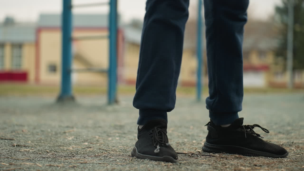 Close-up of legs in motion as someone performs a workout, squatting up and down near an iron pole, a blurred yellow building and a person walking in the background